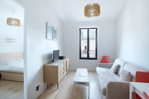 A modern, well-lit room with a bed, a white sofa, a small table, a TV on a wooden cabinet, and a window with a view of a brick building.