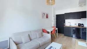 A modern living room with a sofa, a dining table with chairs, a kitchen area with black cabinets and a sink, and a ceiling light fixture. - Apartment Boulogne Facing "les Passages" (Boulogne-Billancourt)