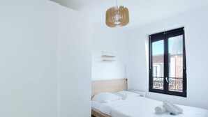 A minimalist bedroom with a bed, a hanging light fixture, a window with a view of buildings, and a small shelf. - Apartment Boulogne Facing "les Passages" (Boulogne-Billancourt)