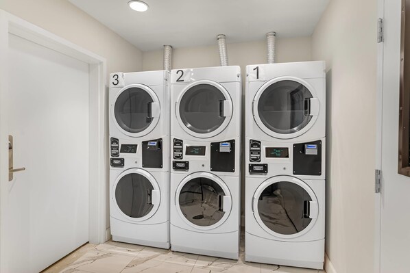 A row of three stacked front-loading washing machines in a laundry room with a white door and tiled floor. - MainStay Suites Chesapeake - Greenbrier (Chesapeake)