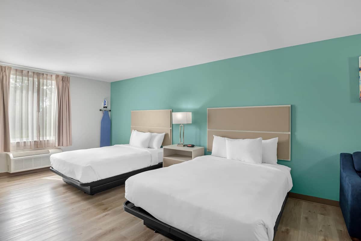 A hotel room with two beds, a window with curtains, a heater, a nightstand with a lamp, and a blue chair.
