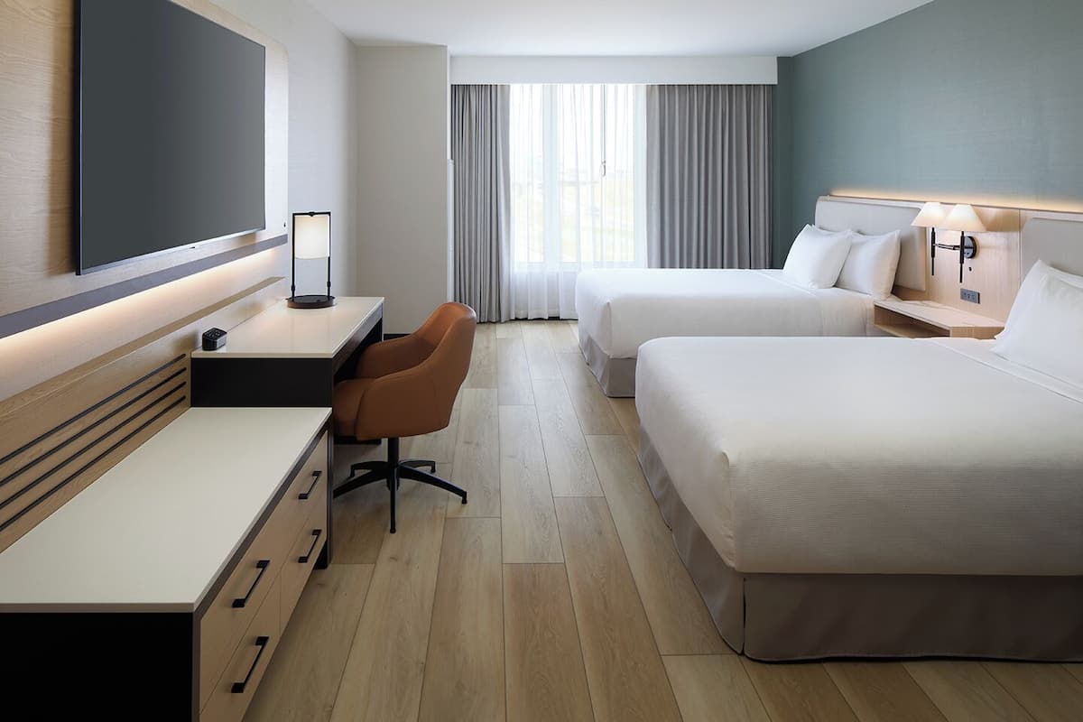Habitación de hotel con escritorio, dos camas, una silla y un ventanal con cortinas.