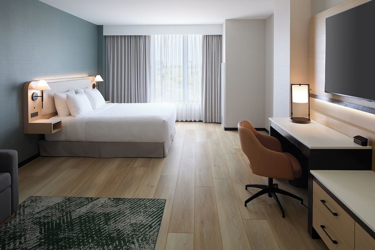 Una habitación de hotel con cama, escritorio, silla y ventana grande con cortinas.
