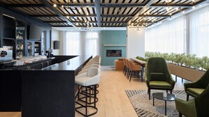 Un área de bar moderna con una barra alargada, diversas opciones de asientos que incluyen taburetes y sillas, una chimenea y un amplio ventanal que proporciona luz natural.
