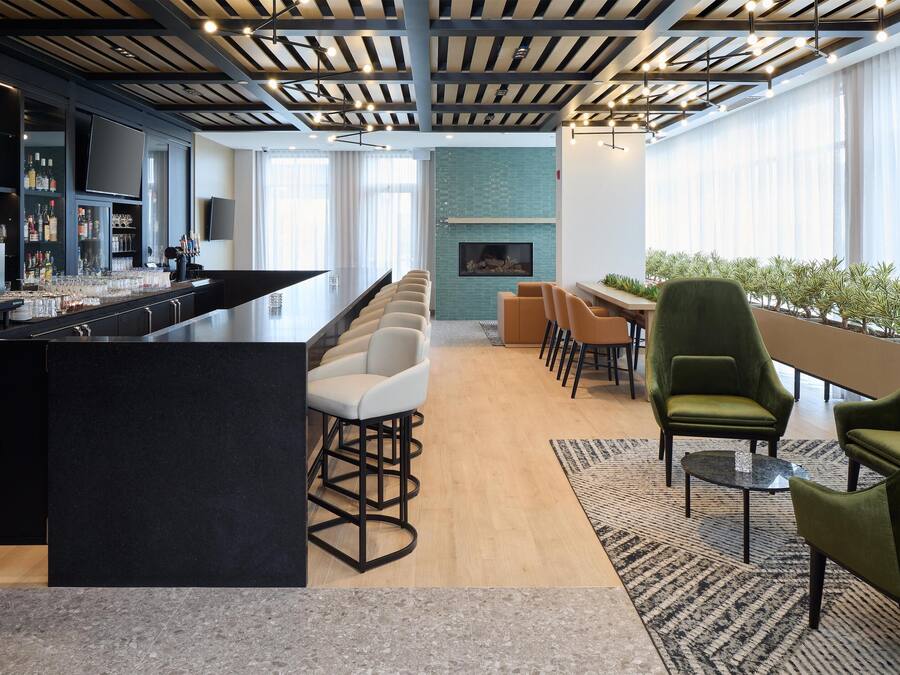 Un espace bar moderne doté d’un long comptoir, de nombreux types de sièges, notamment des tabourets et des chaises de bar, d’une cheminée et d’une grande fenêtre laissant entrer la lumière naturelle.