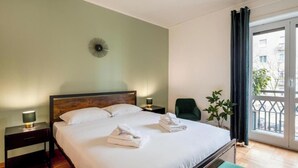 Ein Hotelzimmer mit einem großen Bett, einem Nachttisch mit Lampe, einem Sessel und Blick ins Freie durch ein Fenster.