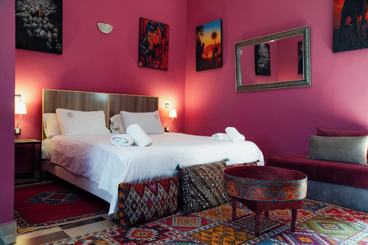 A hotel room with a large bed, two bedside tables with lamps, a red sofa, a patterned rug, and artwork on the walls.