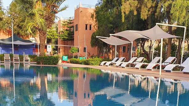 A swimming pool with lounge chairs and umbrellas, surrounded by greenery and a building with a unique architectural style. - Couleurs Berbères (Lalla Takarkoust)