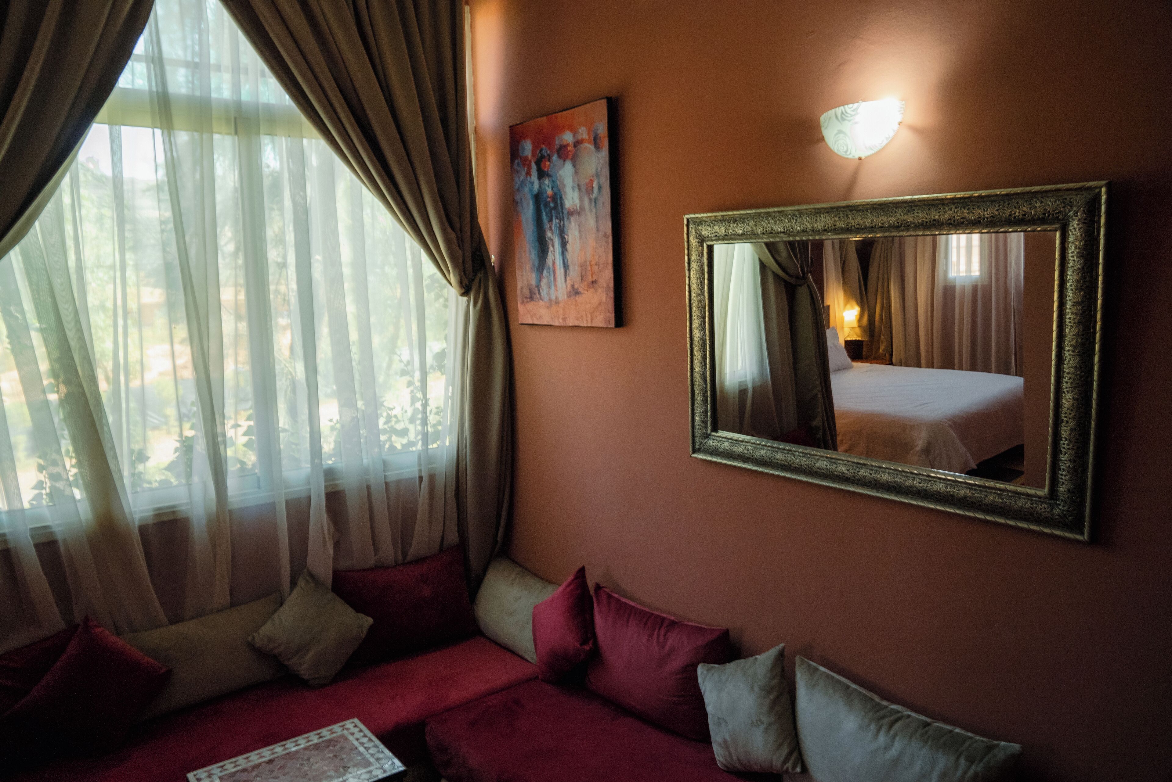 A hotel room with a bed, a large mirror, a painting, and a window with sheer curtains.