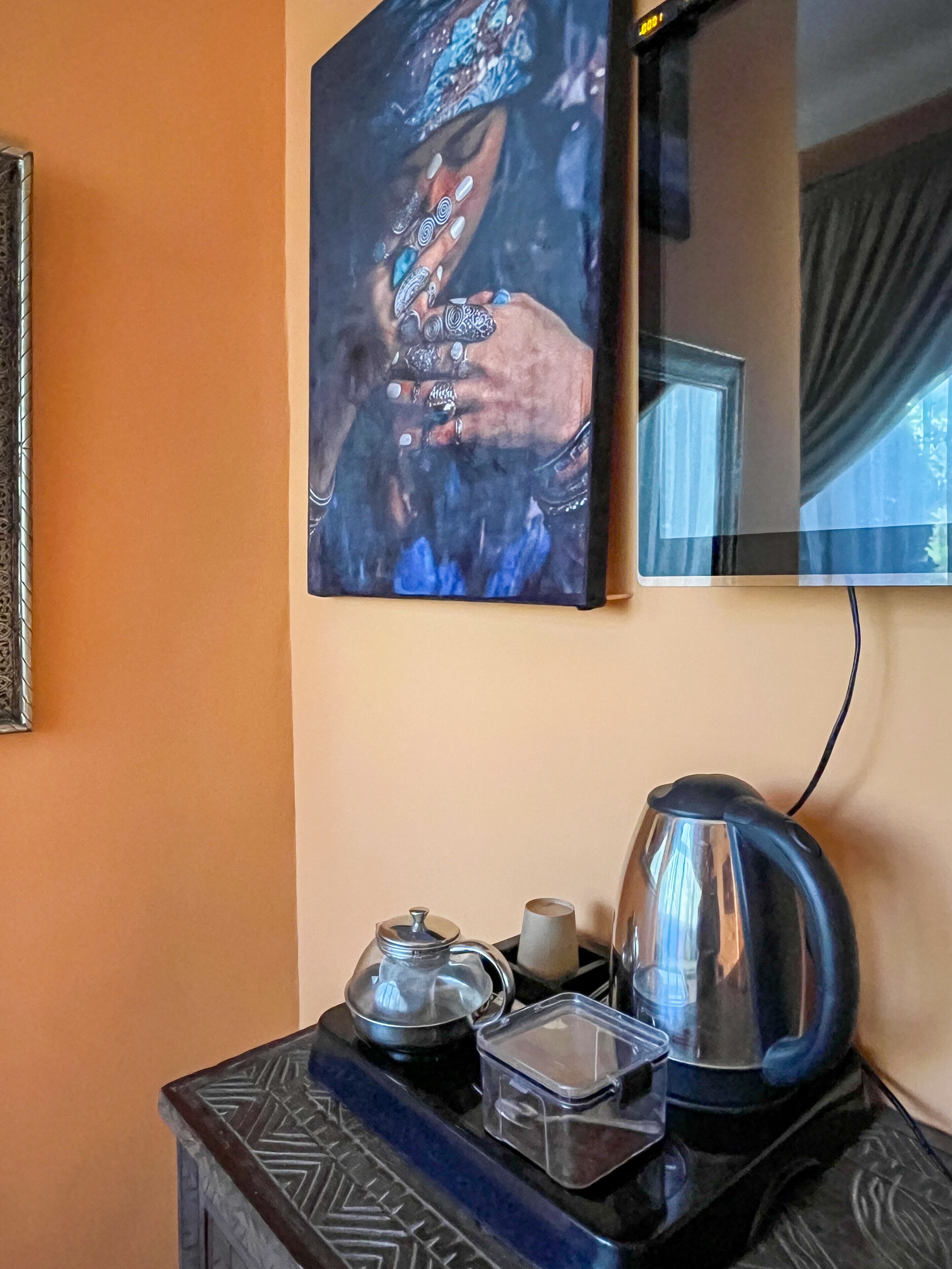 Un cuadro colgado en la pared que muestra manos adornadas con joyas, una cafetera, un hervidor y un recipiente de vidrio sobre una mesa de madera.