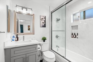 A modern bathroom with a large mirror, a toilet, a sink with a plant, and a glass-enclosed shower area. - Comfy Retreat With Yard Cozy Interiors (San Gabriel)
