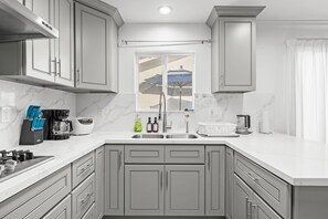 A modern kitchen with grey cabinets, a white countertop, and a window with a view of a beach. - Comfy Retreat With Yard Cozy Interiors (San Gabriel)