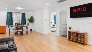 A modern living room with a wooden floor, a dining table with chairs, a sofa, a TV mounted on the wall, and a bookshelf. - Comfy Retreat With Yard Cozy Interiors (San Gabriel)