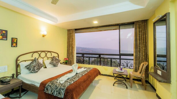 A hotel room with a bed, a table, a chair, and a balcony with a view. - Indira Nature Valley (Mahabaleshwar)