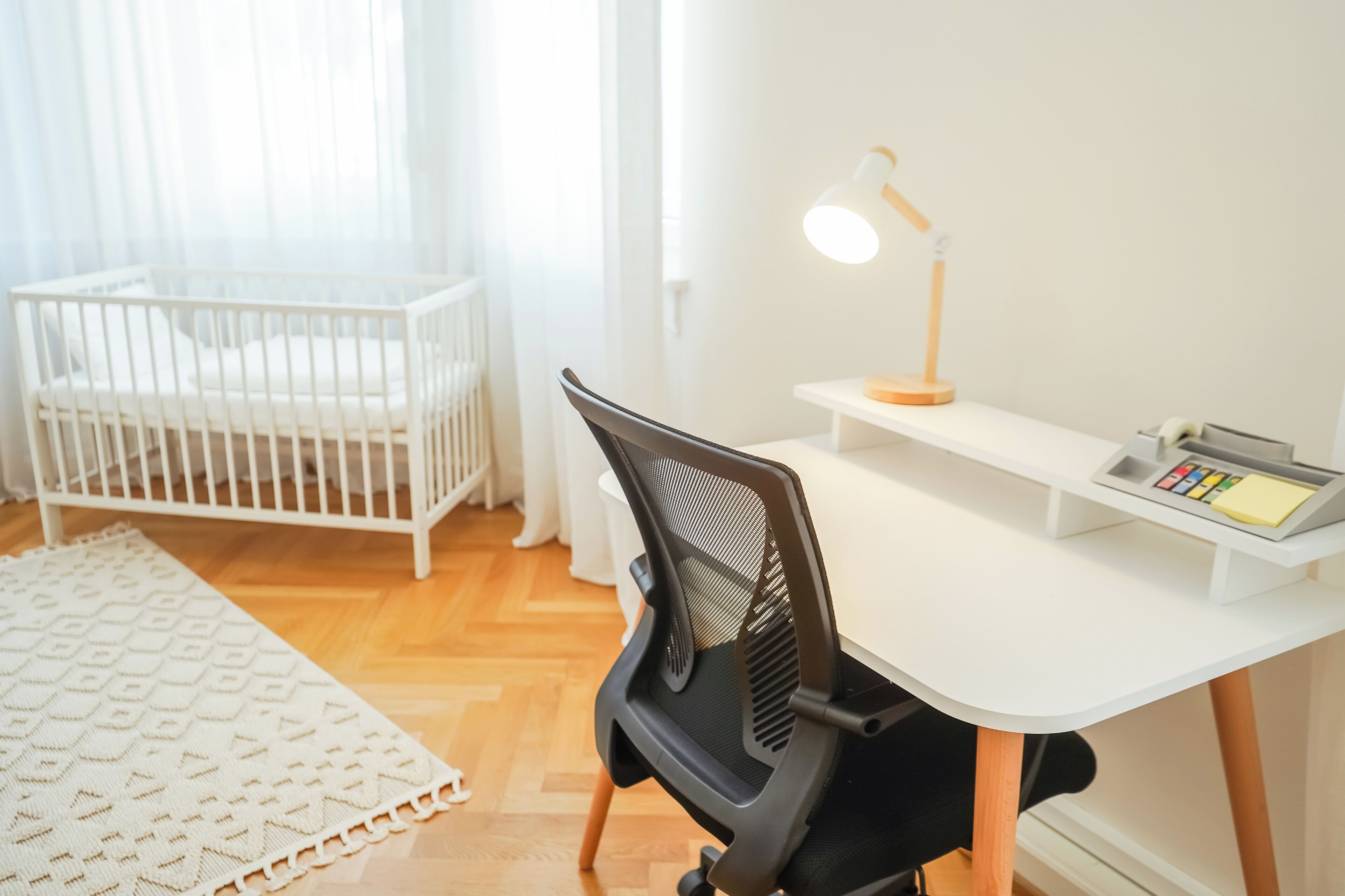 A room with a white crib, a desk with a chair, a lamp, and a control panel.