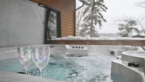 A hot tub with two glasses on the edge, a wooden building, and snow-covered trees in the background. - Manwë | Panoramique | Spa & Jeux by Alchalet (Petite-Rivière-Saint-François)