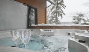 A hot tub with two glasses on the edge, a wooden building, and snow-covered trees in the background.