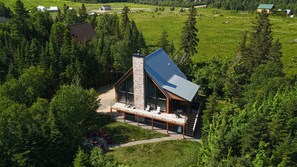 A modern cabin with a large glass facade, a stone chimney, and a solar panel roof, surrounded by dense forest. - Manwë | Panoramique | Spa & Jeux by Alchalet (Petite-Rivière-Saint-François)