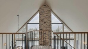 A high-ceilinged room with a stone fireplace, large windows, and a wooden staircase. - Manwë | Panoramique | Spa & Jeux by Alchalet (Petite-Rivière-Saint-François)