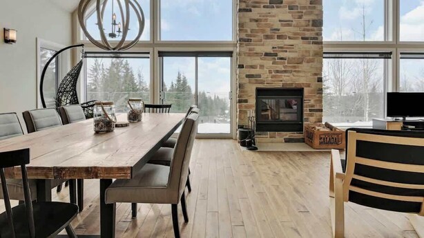 A spacious dining area with a large wooden table, modern chairs, a stone fireplace, and large windows offering a view of the outdoors. - Manwë | Panoramique | Spa & Jeux by Alchalet (Petite-Rivière-Saint-François)