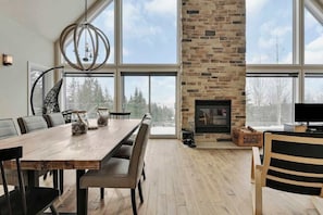 A spacious dining area with a large wooden table, modern chairs, a stone fireplace, and large windows offering a view of the outdoors.