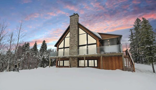 A modern two-story house with a stone chimney, large windows, and a wooden deck.