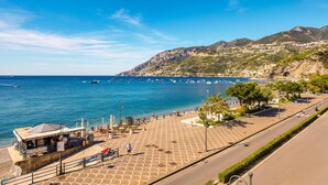 A coastal promenade with a paved walkway, a small kiosk, and a view of the sea and mountains.
