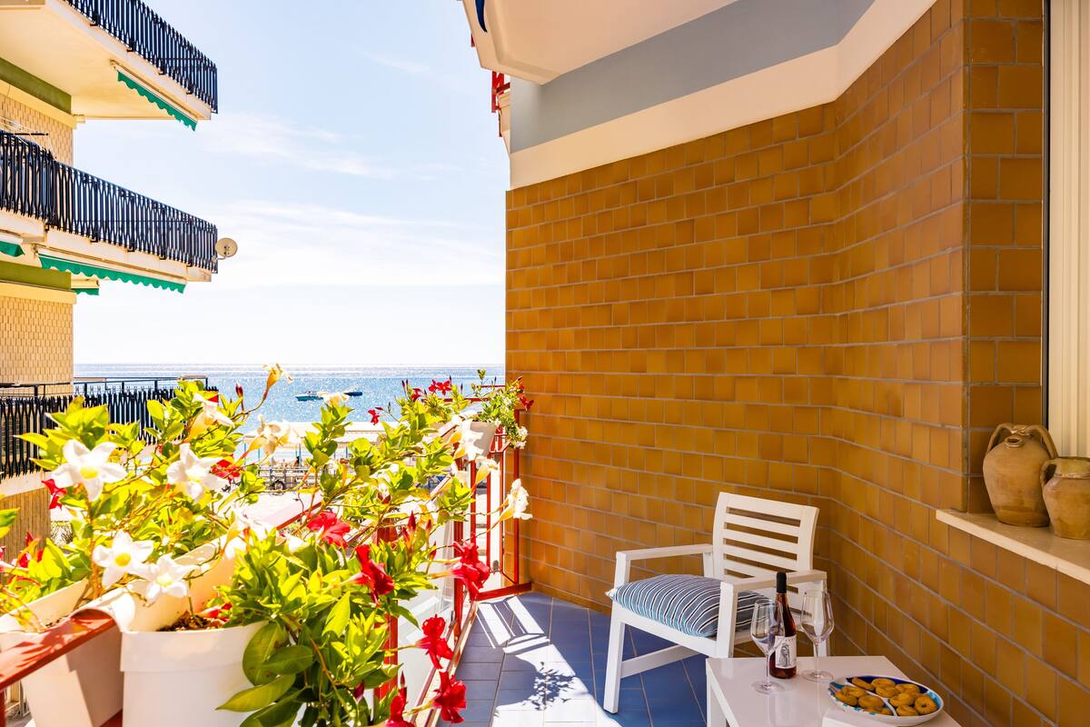Un balcone con vista sul mare, una sedia bianca, un tavolo con una bottiglia e dei bicchieri, e piante in vaso.