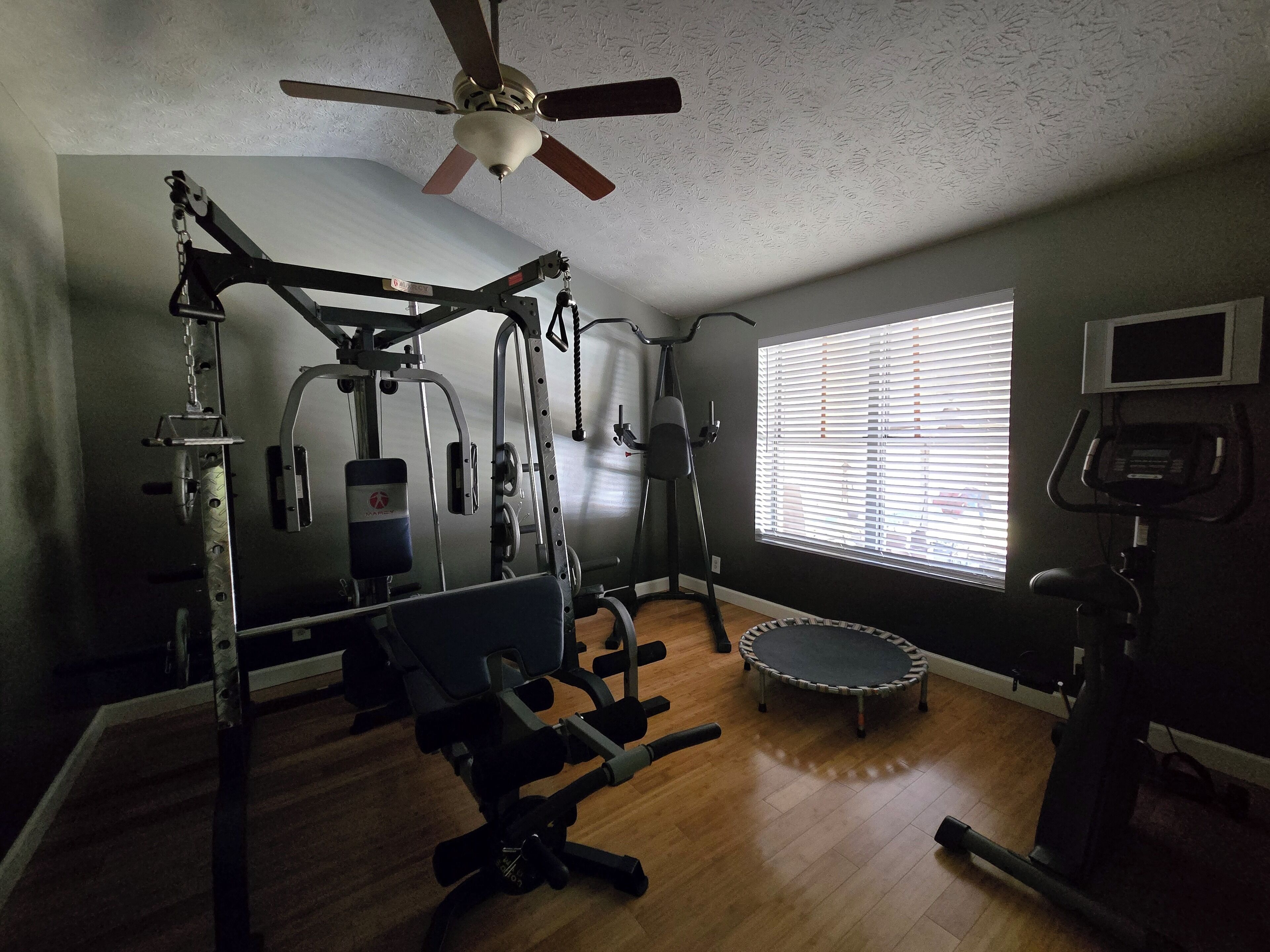 A home gym with various exercise equipment, a ceiling fan, and a window with blinds.