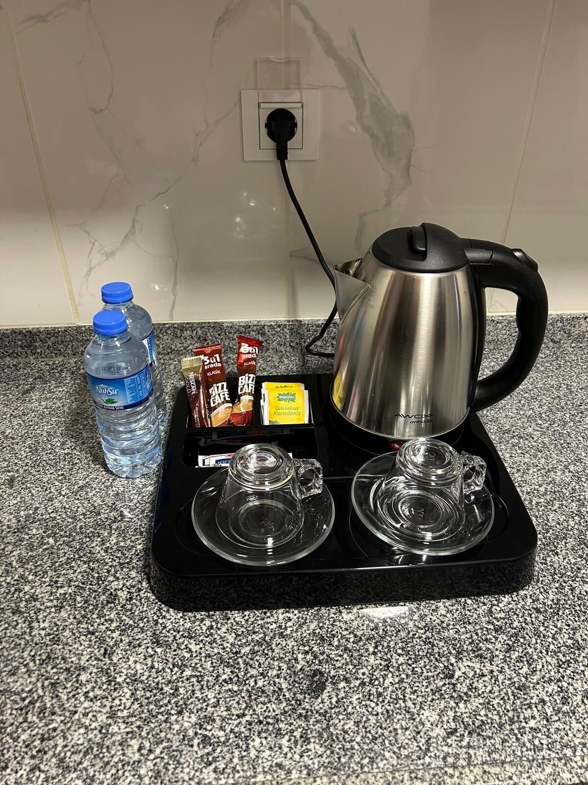 A kitchen counter with a kettle, two glass cups, a water bottle, and some food items.