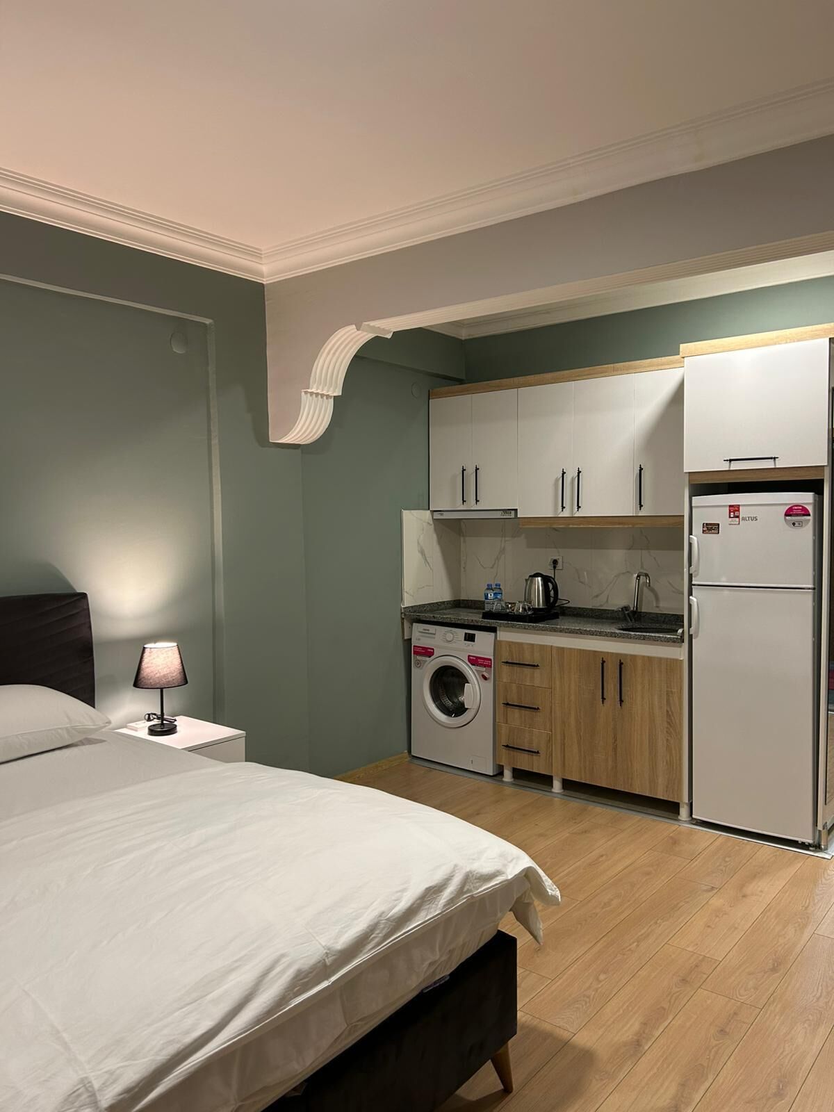 Ein gemütliches Schlafzimmer mit Bett, Nachttisch, Lampe und einer Waschmaschine in einer Küchenzeile mit Schränken und Kühlschrank.