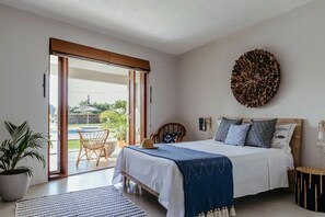 A bedroom with a bed, a large circular wall decoration, a potted plant, and a view through a sliding door to an outdoor area with a table and chairs.