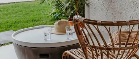 A wicker chair and a round table with a hat and glasses on it, placed on a patio with a large potted plant and a view of a road and greenery.
