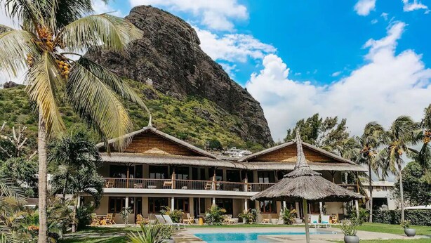 A resort with a pool, thatched-roof structures, and a mountain backdrop. - Boutik Le Morne Holiday Apartments (Le Morne)