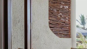 A room with a round wooden table and two wooden chairs, a hat on the table, and a textured wall with a woven panel. - Boutik Le Morne Holiday Apartments (Le Morne)