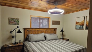Ein Schlafzimmer mit Holzboden, einem Bett mit gestreifter Bettwäsche, zwei Nachttischen mit Lampen, einem Fenster mit Jalousien und Wandbildern.