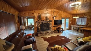 Ein gemütliches Blockhaus-Interieur mit Kamin aus Stein, Ledersofas, einem Essbereich und einem an der Wand montierten Fernseher.
