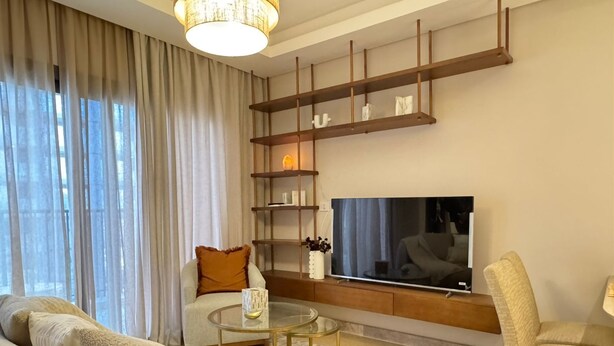 A modern living room with a sofa, armchairs, a coffee table, a TV mounted on a wooden shelf, and a large window with sheer curtains. - Mynt stay hotel apartment - zed towers (Sheikh Zayed City)