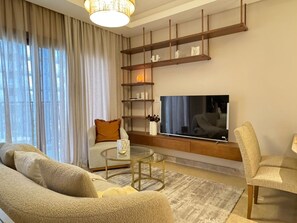Un soggiorno moderno con divano, poltrone, tavolino, TV fissata a una mensola di legno e un'ampia finestra con tende leggere.