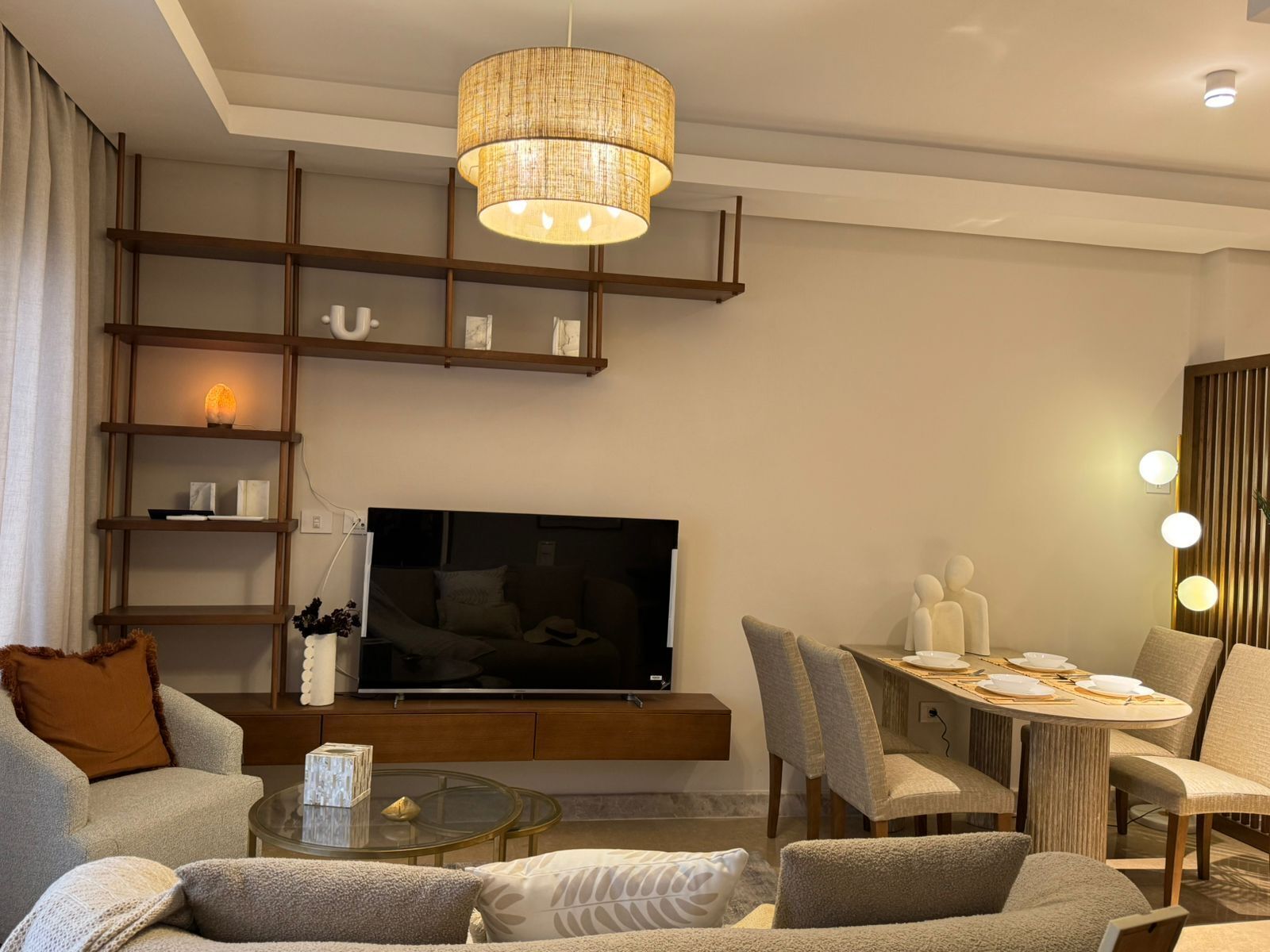 A modern living room with a flat-screen TV, comfortable seating, a glass coffee table, and a dining area with a wooden table and chairs.