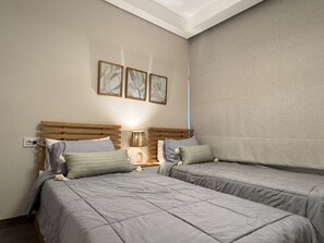Two single beds with wooden headboards and grey bedding, a nightstand with a lamp, and framed artworks adorn this serene bedroom.