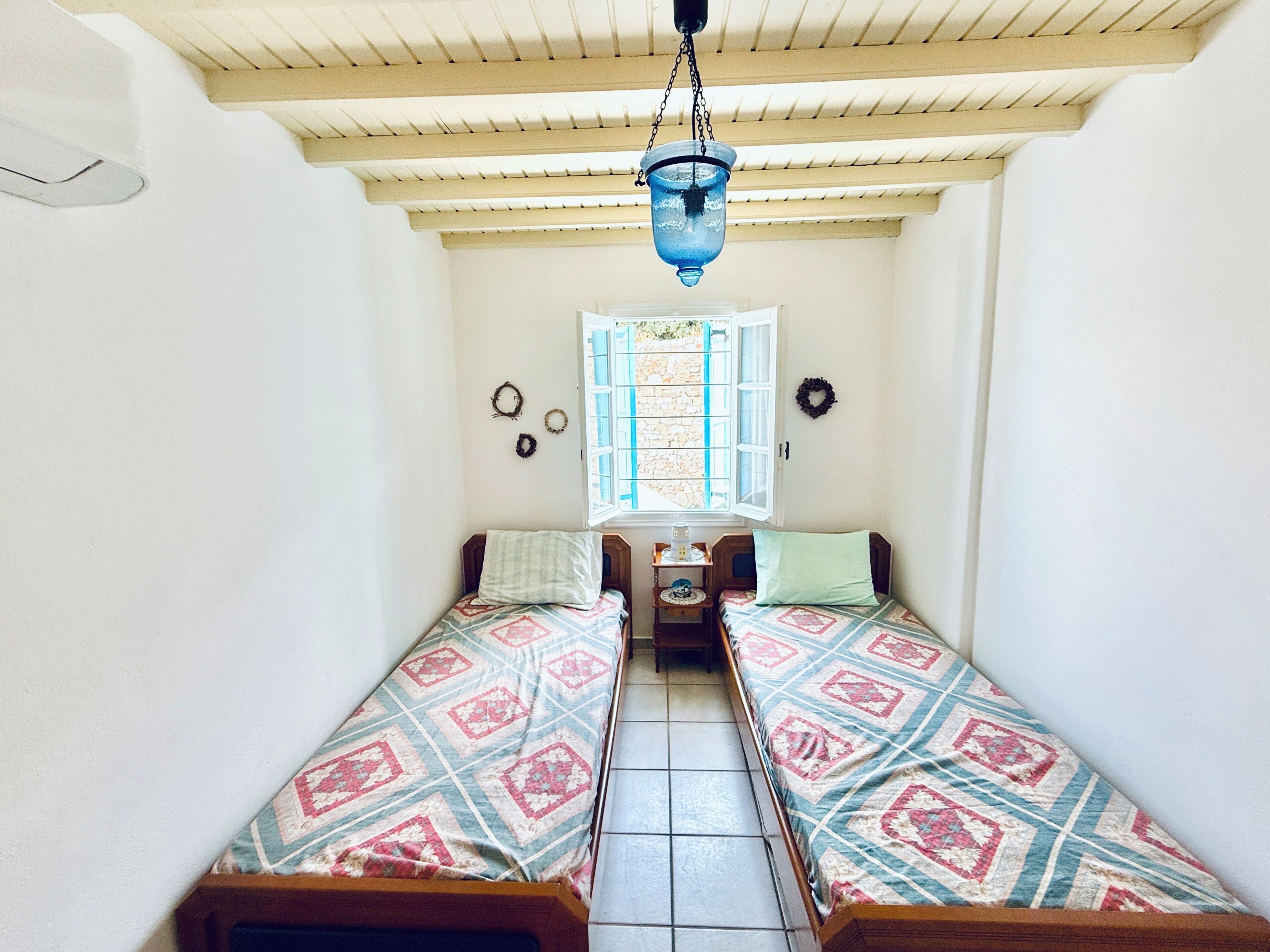 A small, cozy room with two single beds, a window with shutters, a hanging light fixture, and a small table with a lamp.