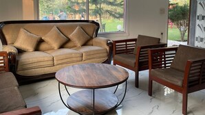 A spacious living room with a large window, a sofa, two armchairs, and a coffee table. - Luxsugero Farms (Faridabad)