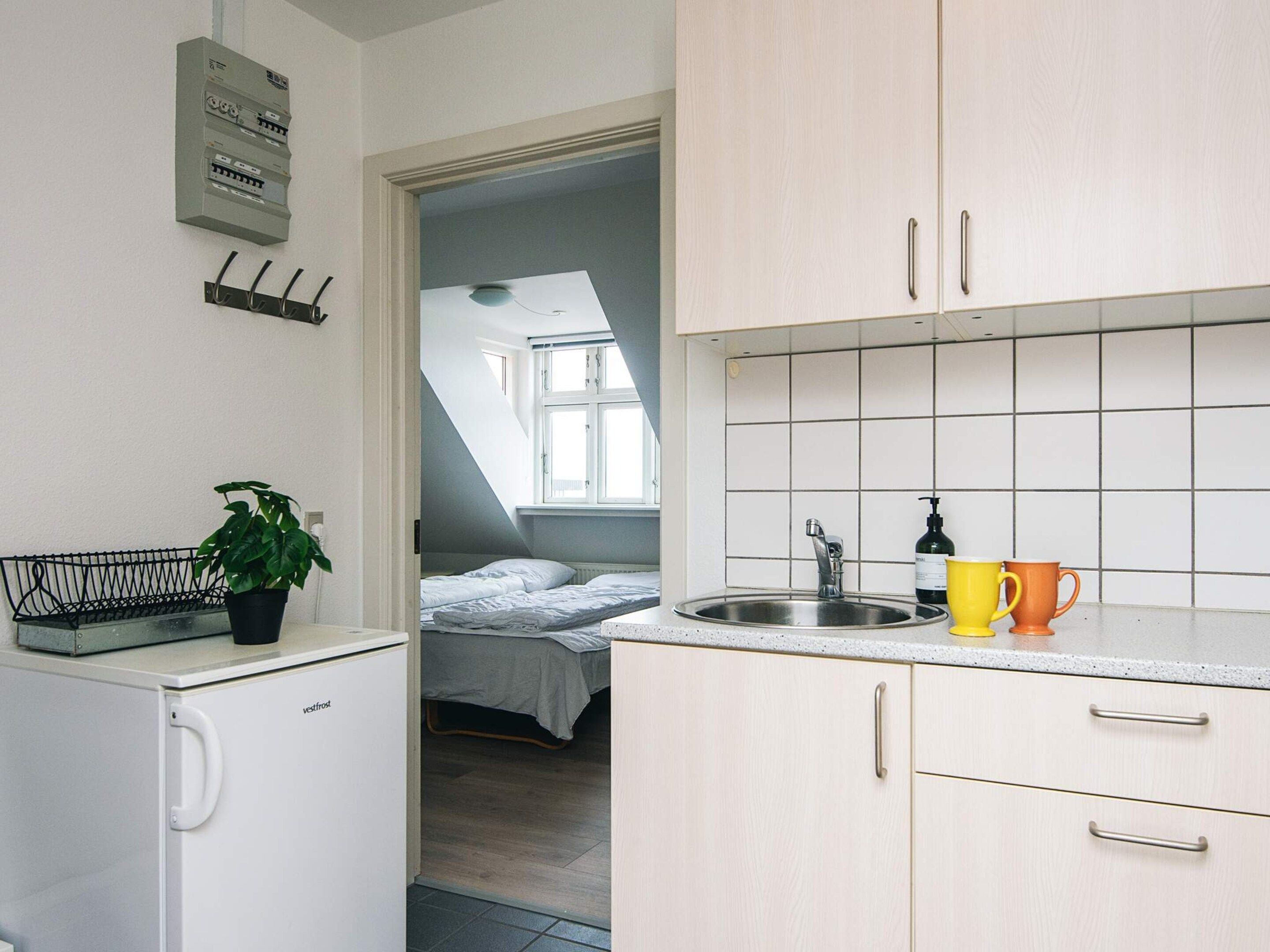 Une cuisine compacte équipée d’un réfrigérateur encastré, d’un évier et d’un plan de travail. On peut y voir une petite plante posée sur le réfrigérateur et une fenêtre au-dessus de l’évier.