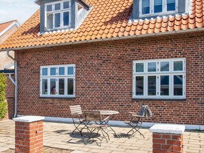 A two-story brick house with a tiled roof, two windows with shutters, and a patio with a table and chairs. - 2 Person Holiday Home in Ringkobing (Ringkobing)
