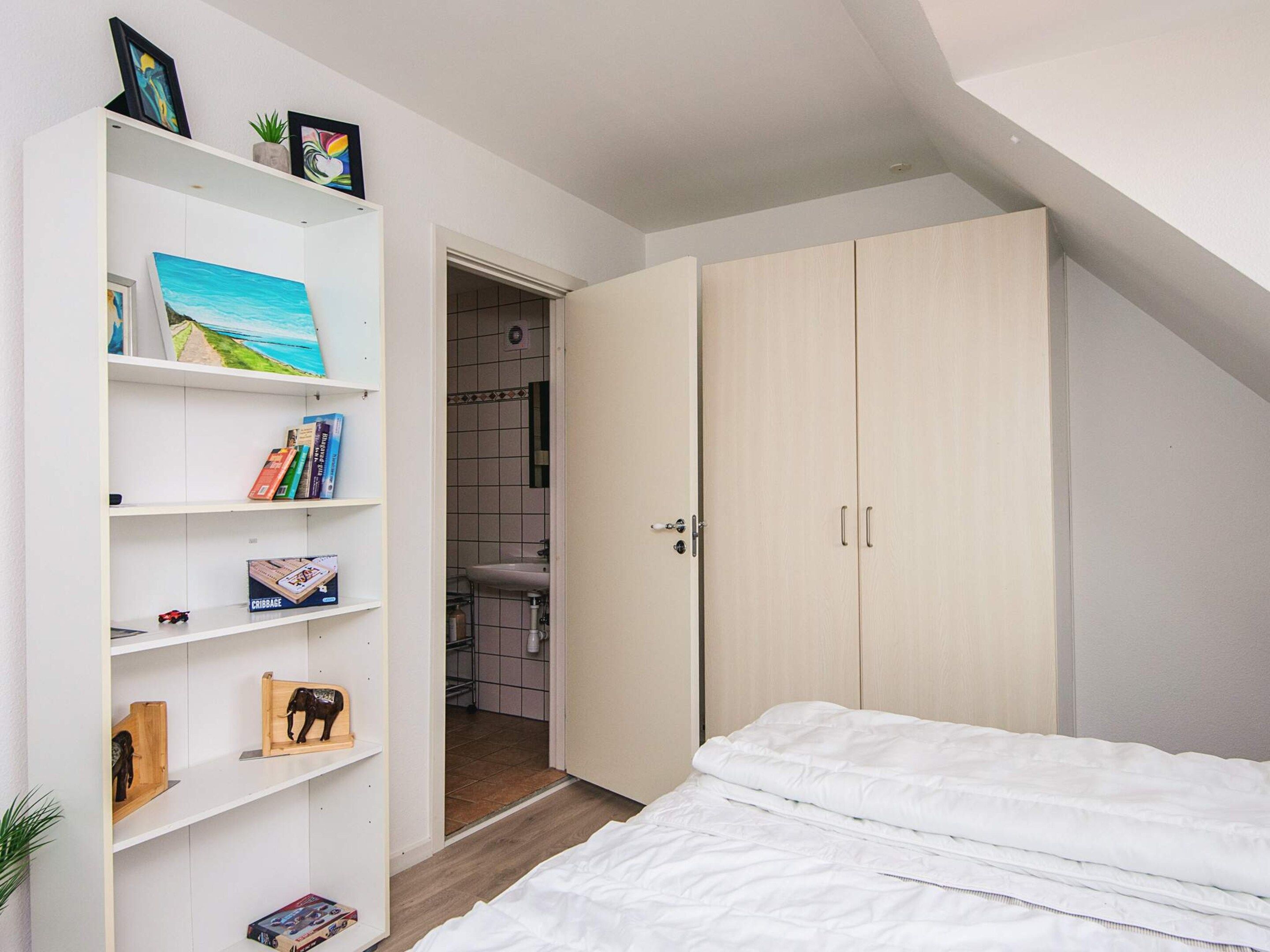 A bedroom with a bed, a white wardrobe, and a bookshelf with various items.