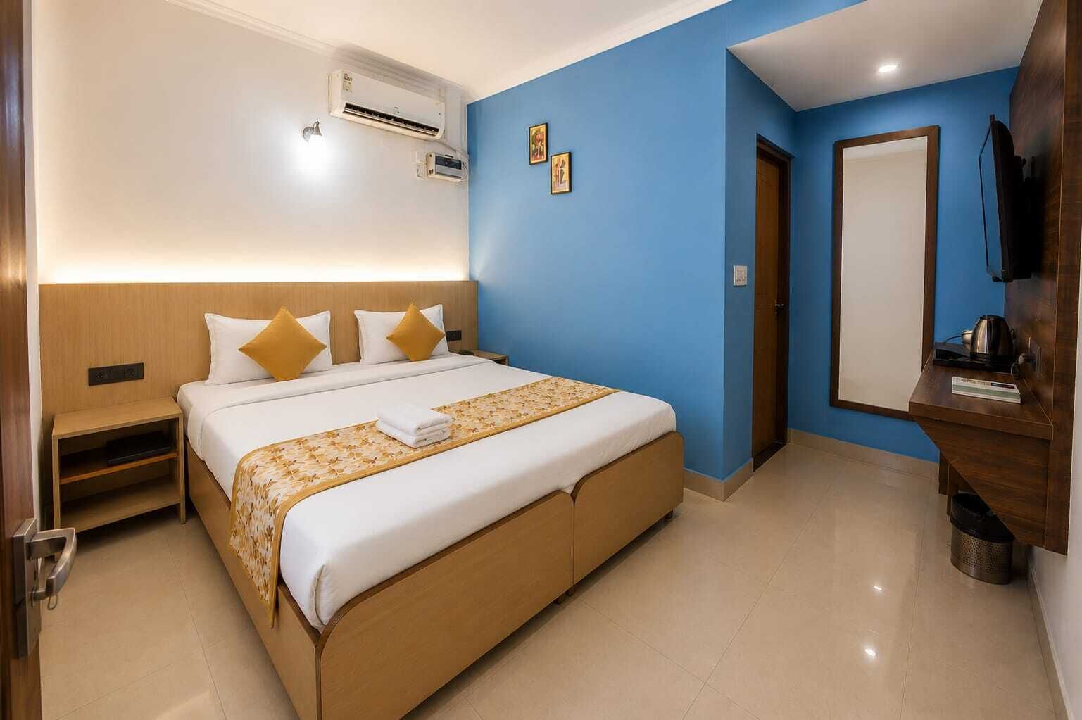 A hotel room with a bed, bedside tables, an air conditioning unit, a television, and a door.