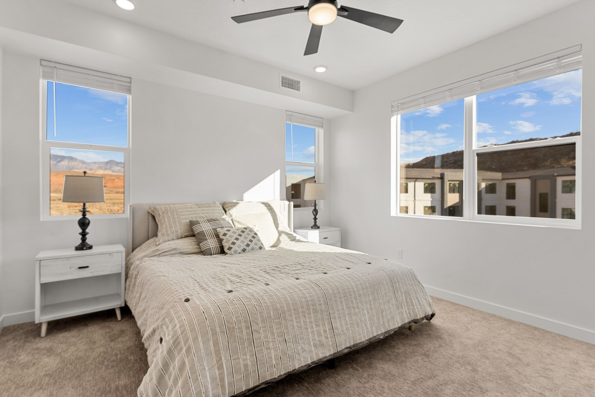 A bedroom with a bed, two nightstands, a ceiling fan, and large windows with views of a desert landscape and buildings.