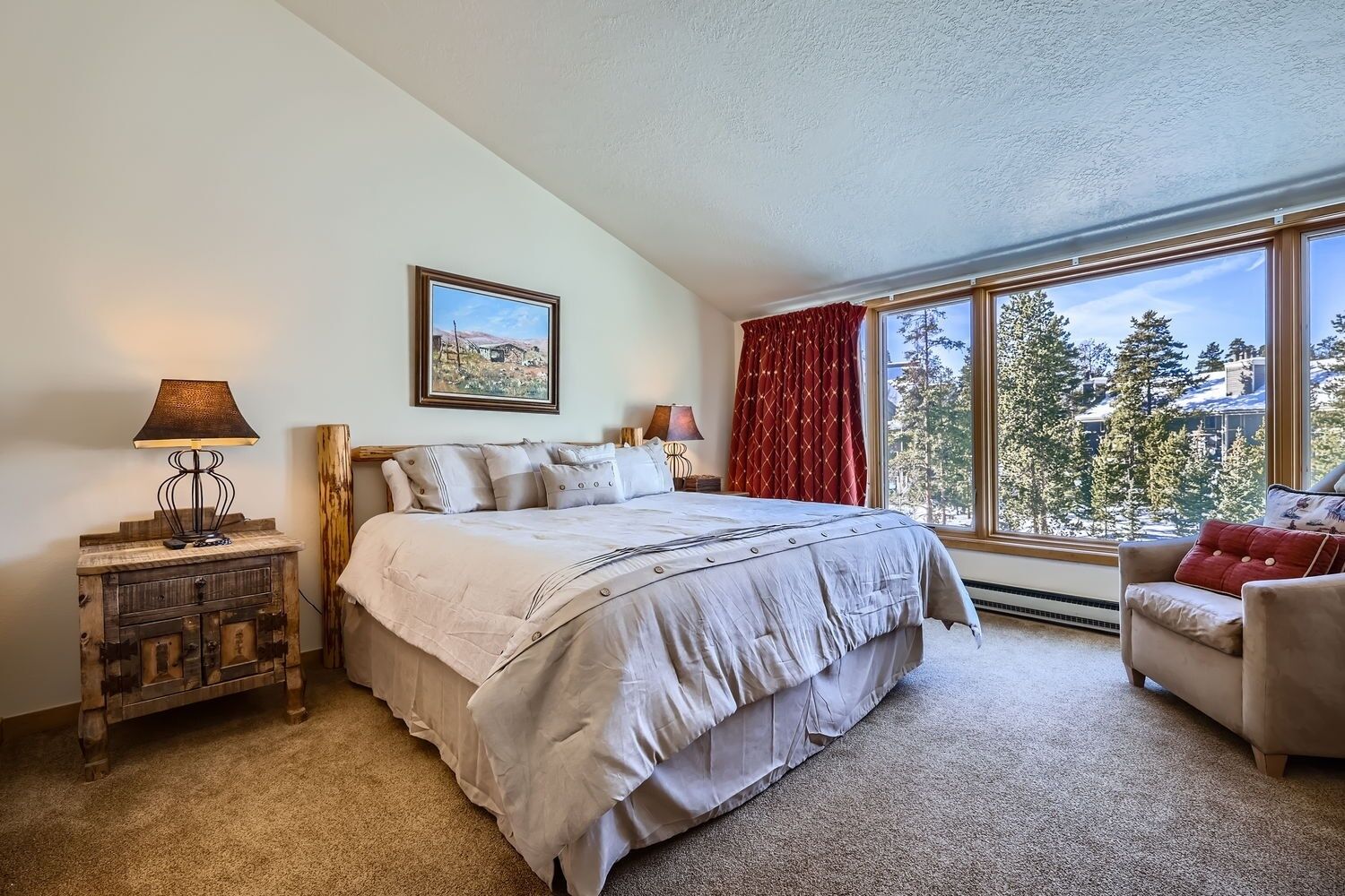 Une chambre à coucher avec un grand lit, une table de chevet, une lampe, une chaise, une fenêtre donnant sur des arbres et un tableau accroché au mur.