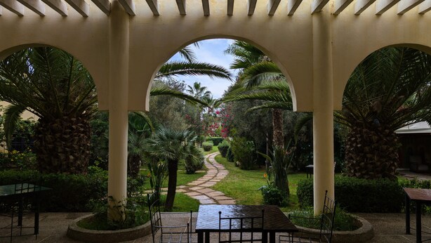 A covered garden pathway with palm trees and a stone path leading to a seating area with tables and chairs. - Les Cypres Hotel Restaurant  (Moulay Abdallah)